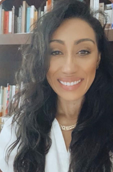 Headshot of Scherly Virgll in a white shirt with long dark hair and a bookshelf in the background. 