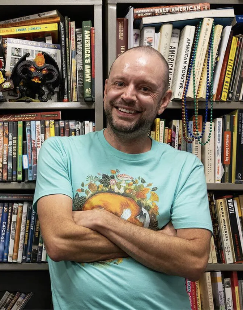 Dr. Chester standing with arms folded and smiling in front of grey bookshelves filled with colorful books, wearing a light blue shirt. 